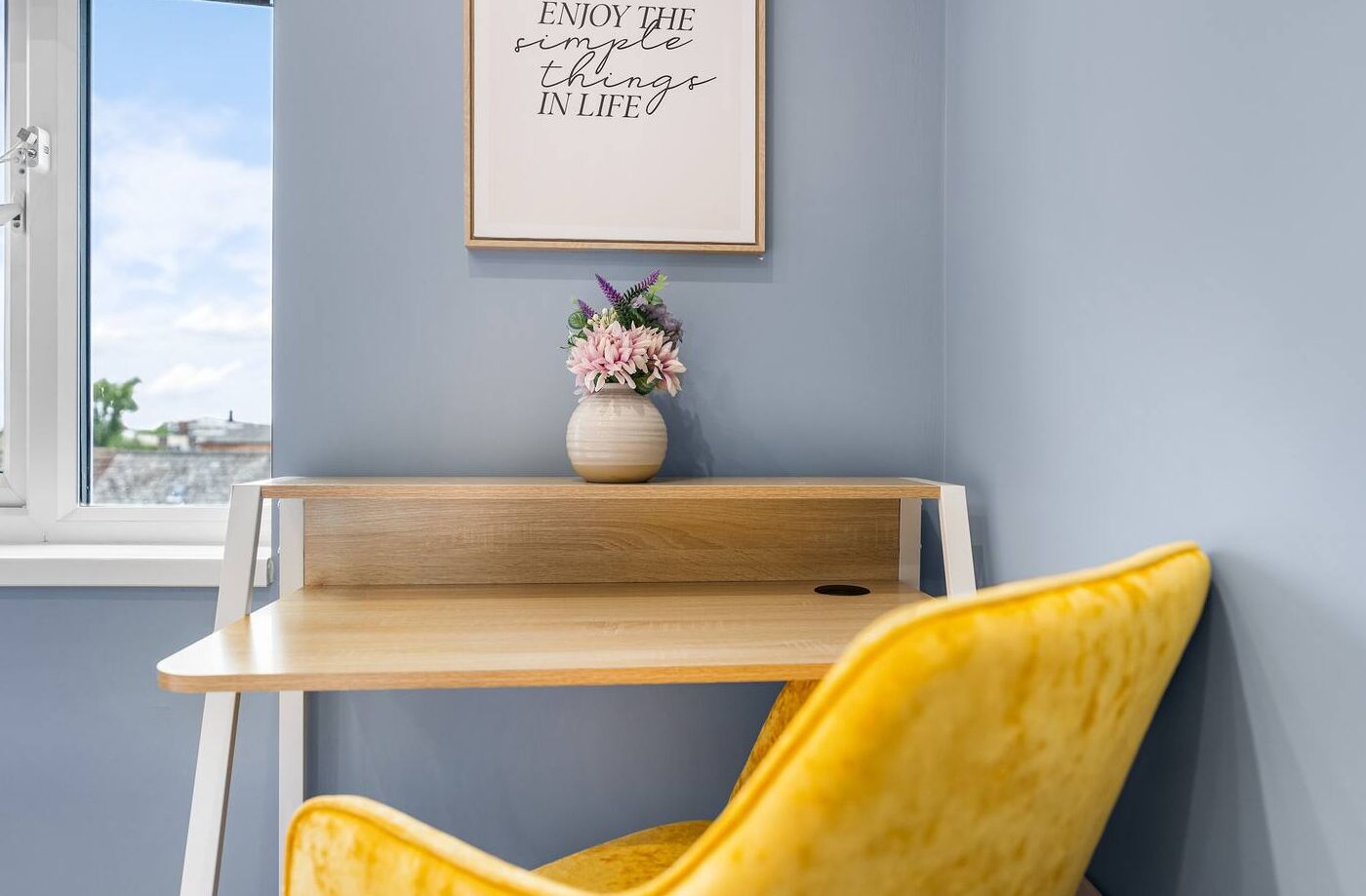Quarto com escrivaninha de madeira, cadeira amarela, um vaso com flores e uma moldura com uma citação inspiradora na parede.