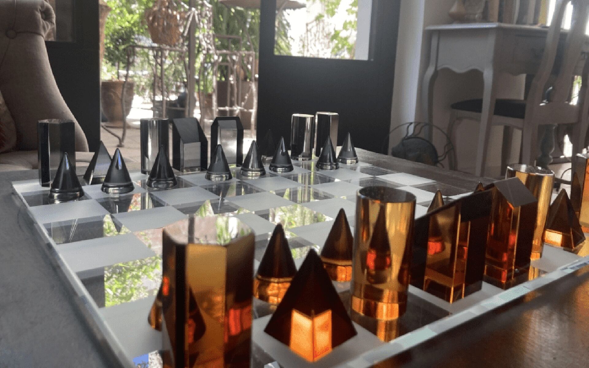 A chessboard with variously shaped and colored pieces, set on a reflective surface in a room with a window and furniture in the background.