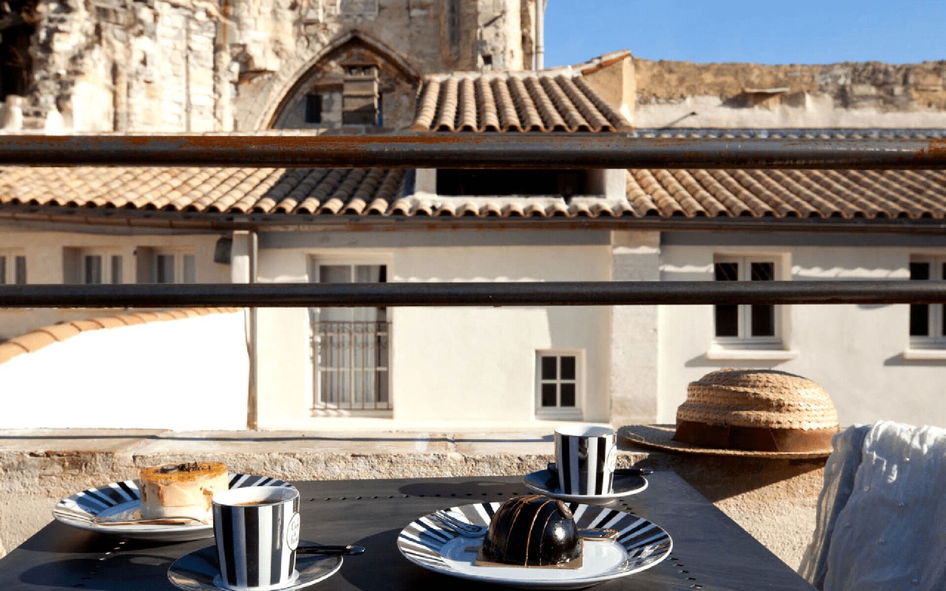 A table with plates, cups, and a basket, set against a building with tiled roofs and a stone wall.