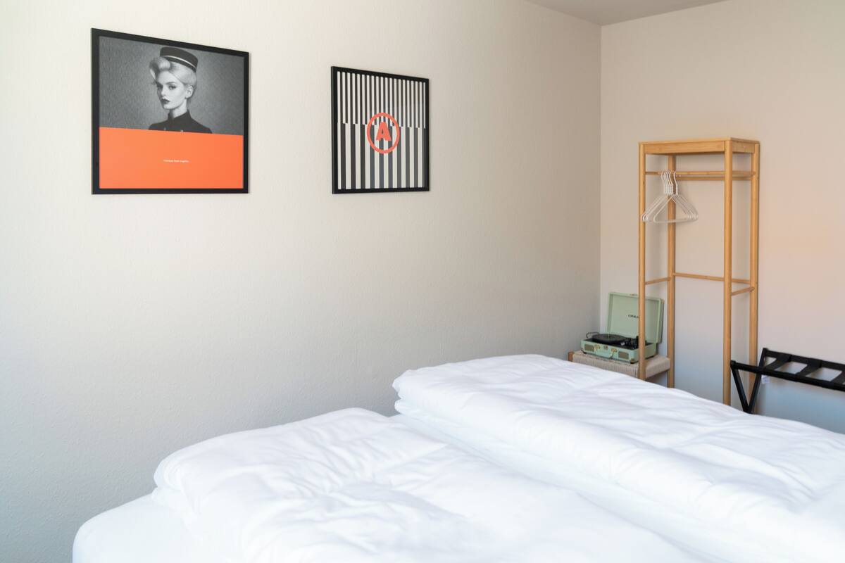 A hotel room with a neatly made bed, a wooden wardrobe, and two framed artworks on the wall.