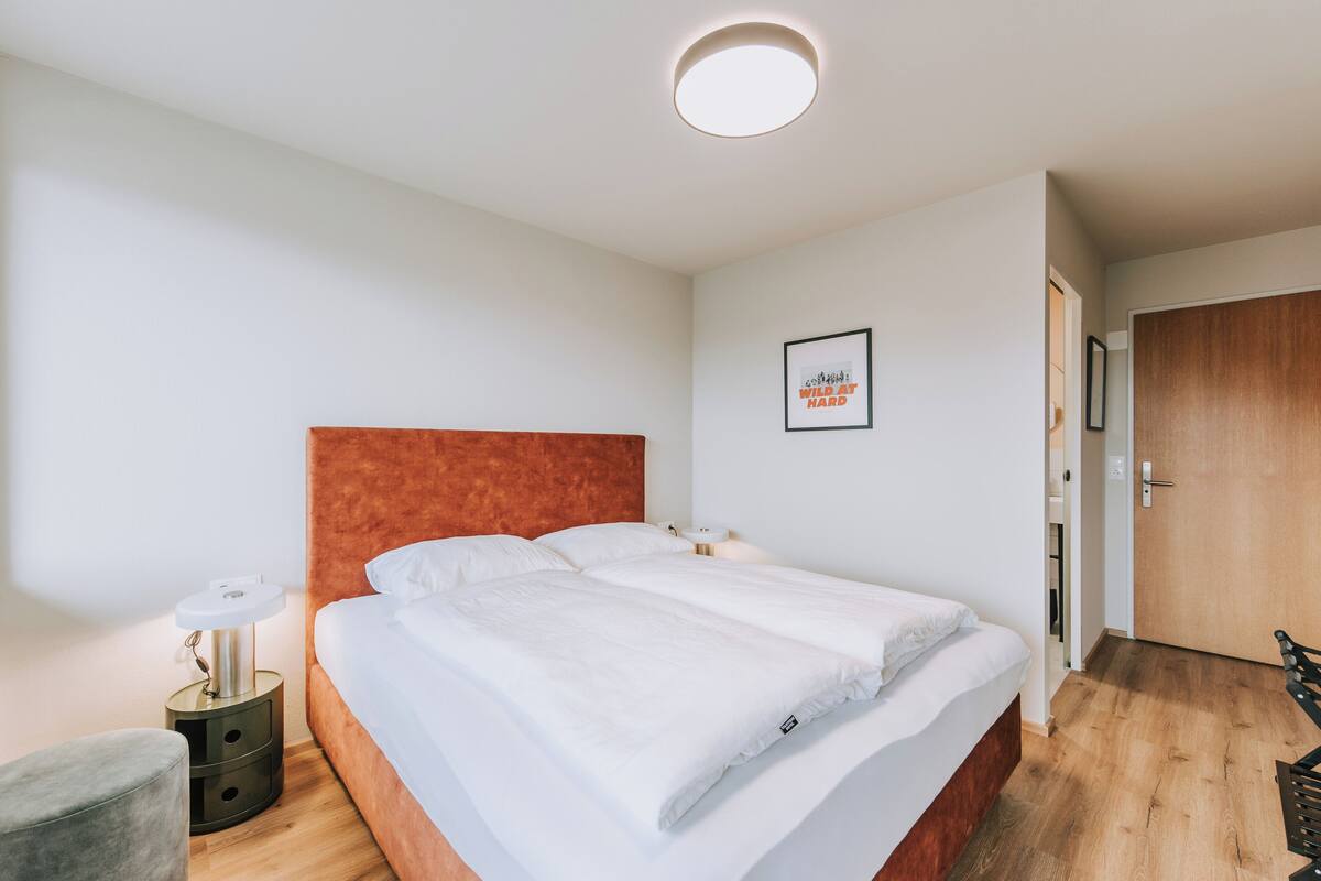 A double bed with white linens, a round ceiling light, a small bedside table with a lamp, and a framed picture on the wall.