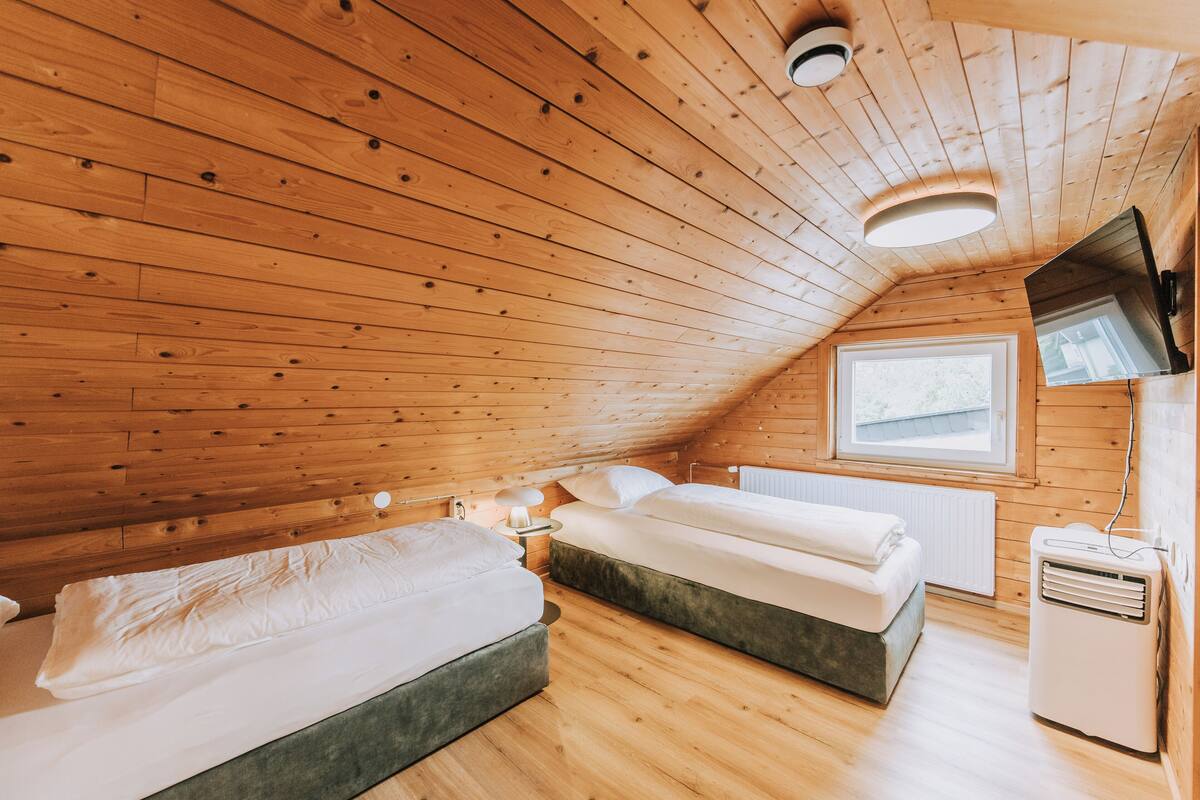 A wooden-paneled room with two beds, a window, a radiator, and a TV mounted on the wall.
