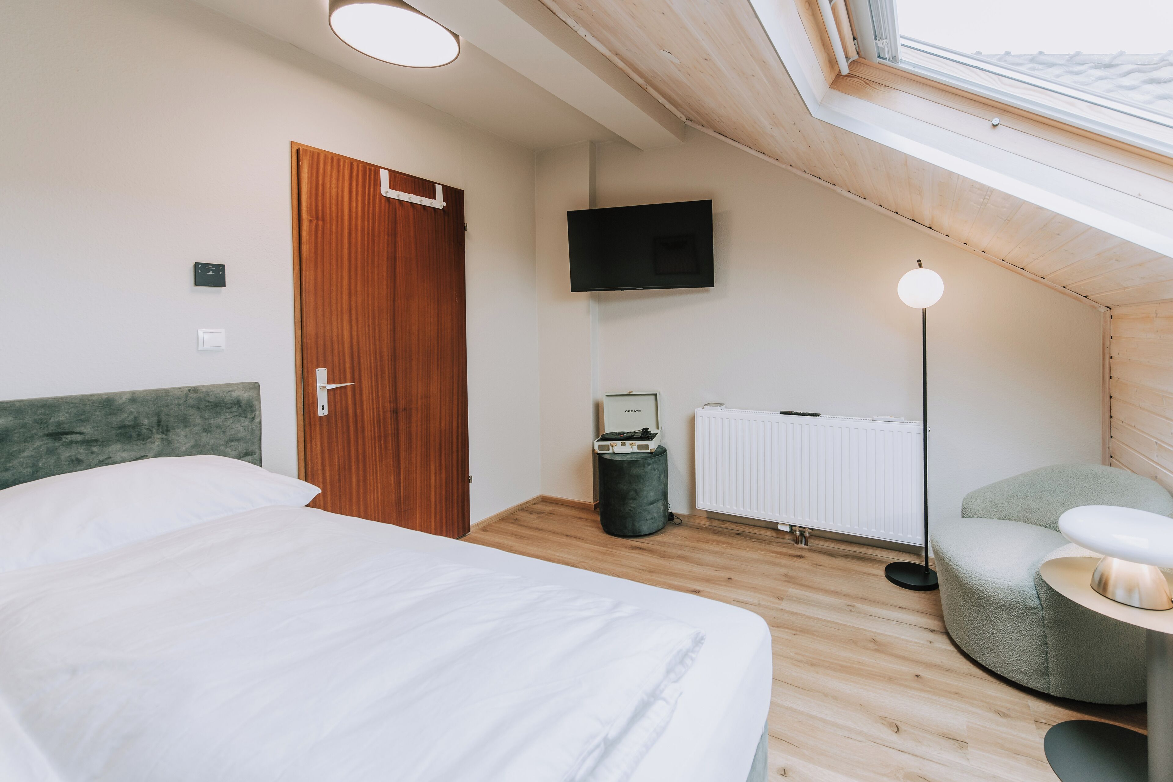 A modern bedroom with a large bed, a wooden door, a flat-screen TV, a radiator, and a skylight.