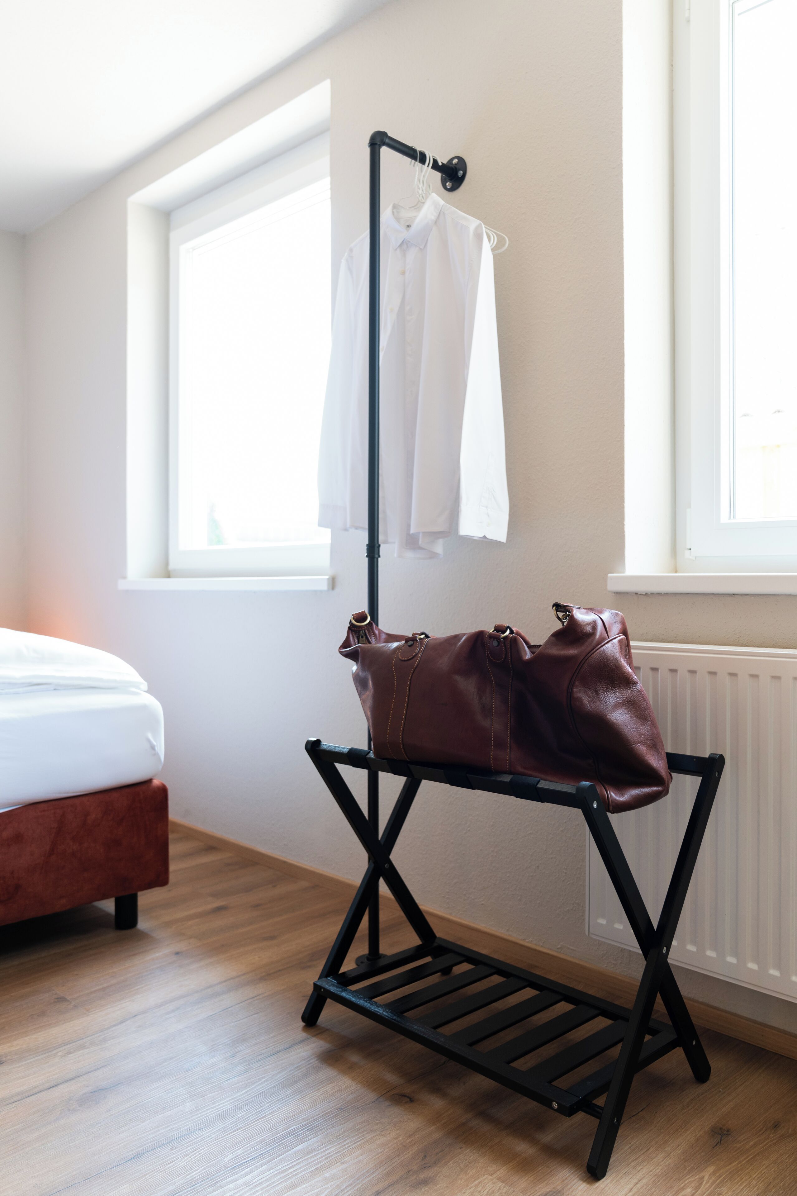 A room with a bed, a radiator, a window, and a black metal drying rack with a brown leather bag.