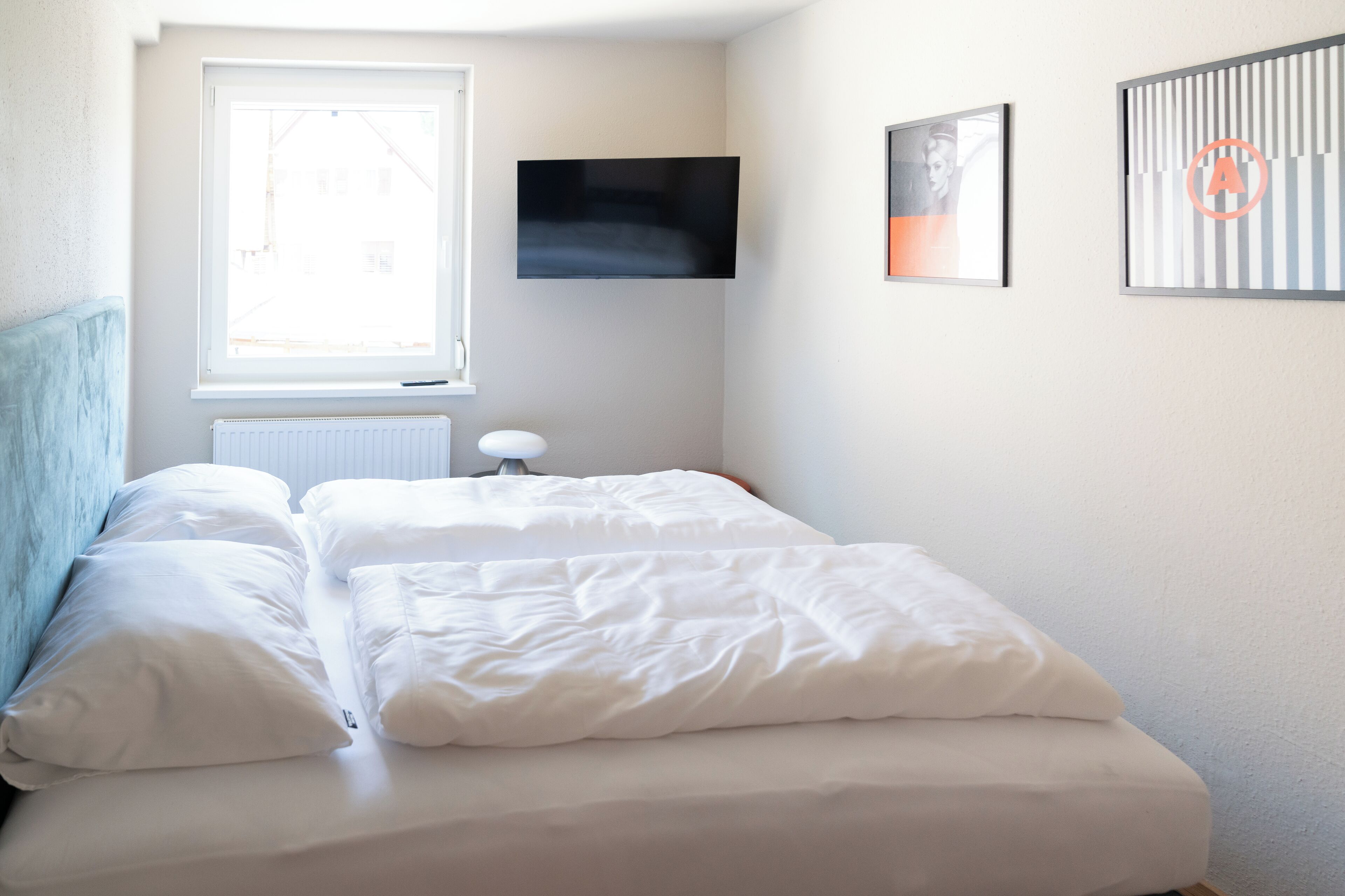 A neatly made bed with white linens in a room with a window, a radiator, and a flat-screen TV mounted on the wall.