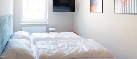 A neatly made bed with white linens in a room with a window, a radiator, and a flat-screen TV mounted on the wall.