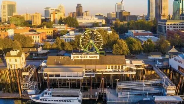 A waterfront building with a Ferris wheel, boats docked in front, and a city skyline in the background. - Barbiecore Dream Pad in Sacramento! (Sacramento)