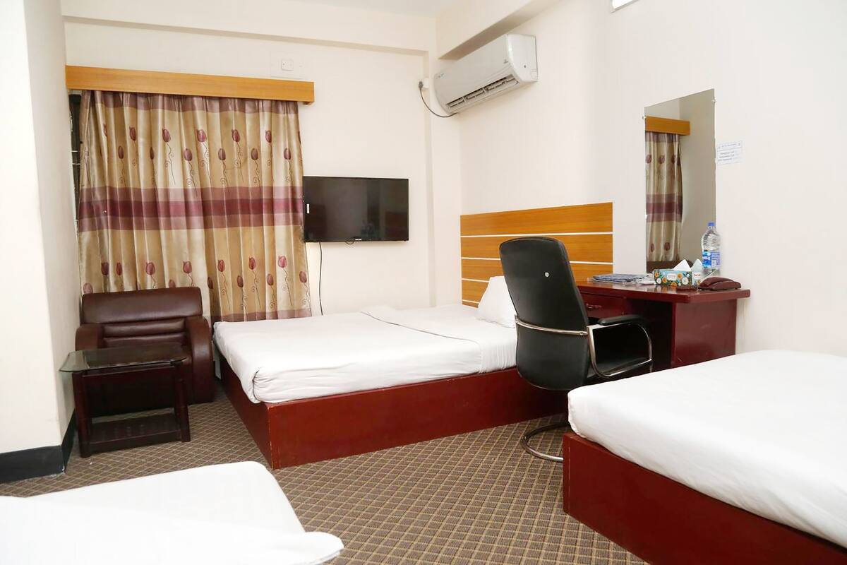 A hotel room with two beds, a desk, a chair, a TV, and an air conditioning unit.