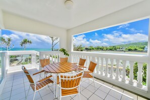 Ein Balkon mit weißer Brüstung, einem runden Holztisch mit Stühlen und Blick auf Meer und Grünflächen.