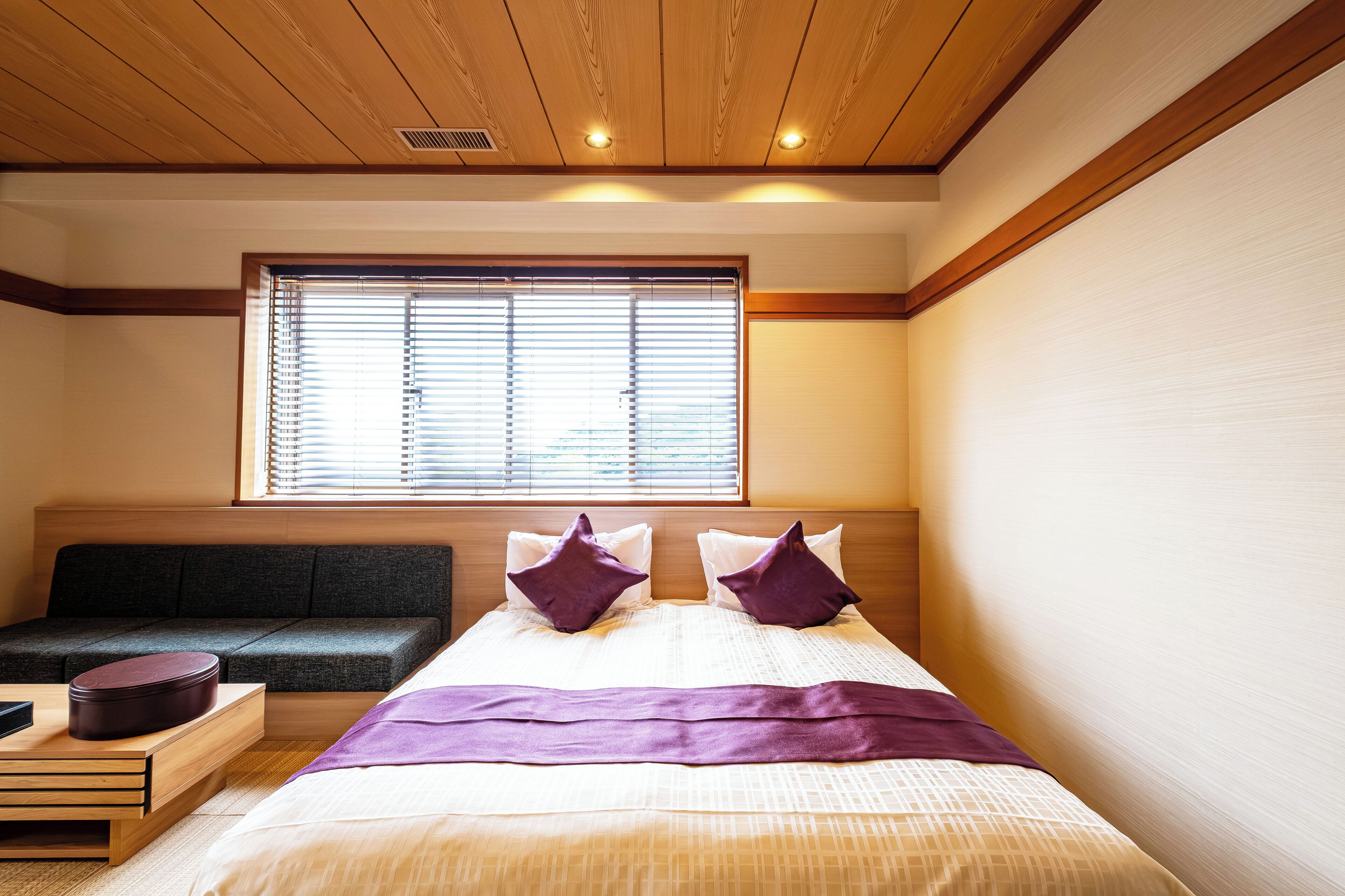 A hotel room with a bed, a sofa, a window with blinds, and a wooden ceiling.