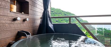 A wooden spa area with a hot tub, a wooden shelf with bottles, and a view of a green hillside.