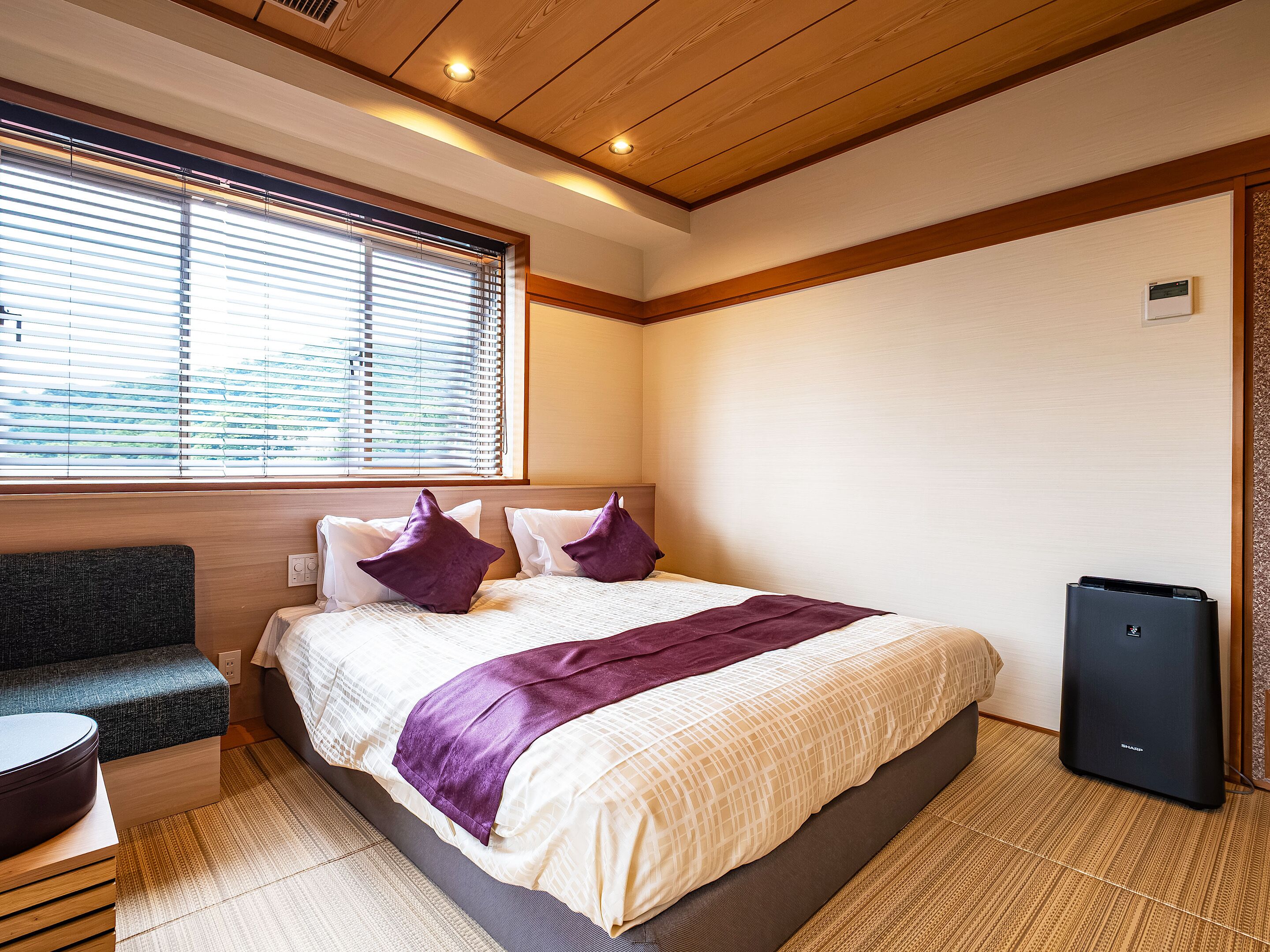 A hotel room with a bed, a bench, a heater, and a window with blinds.