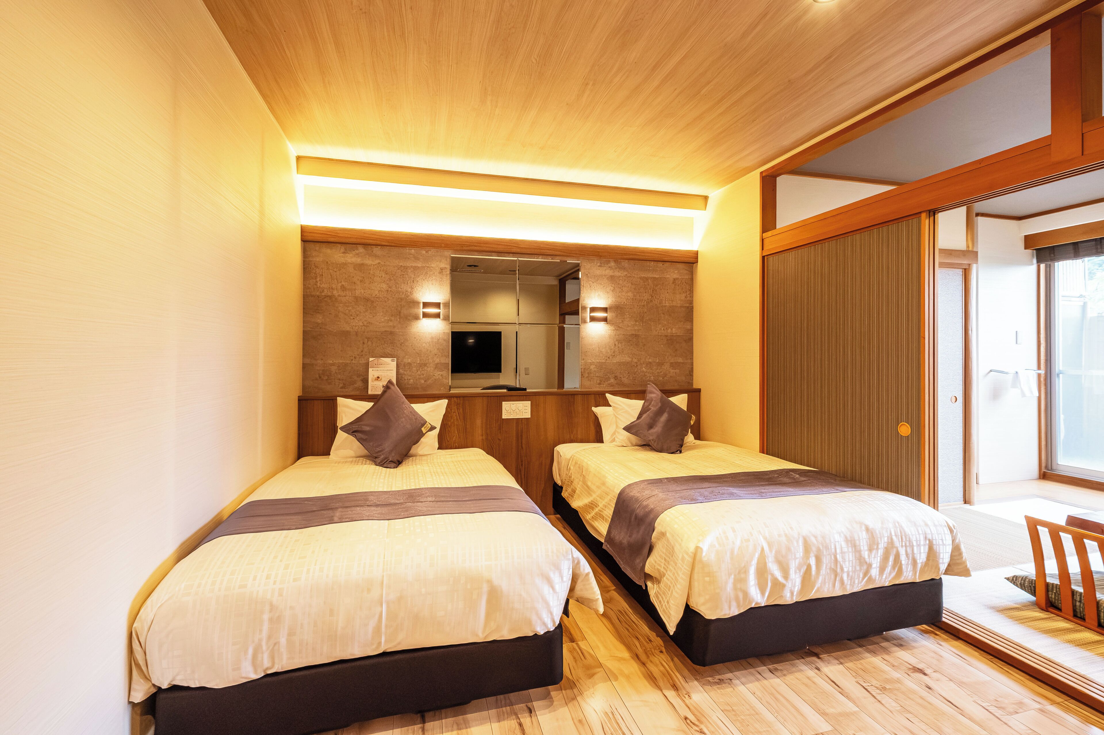 A hotel room with two beds, wooden flooring, a built-in TV, and a sliding door to a balcony.
