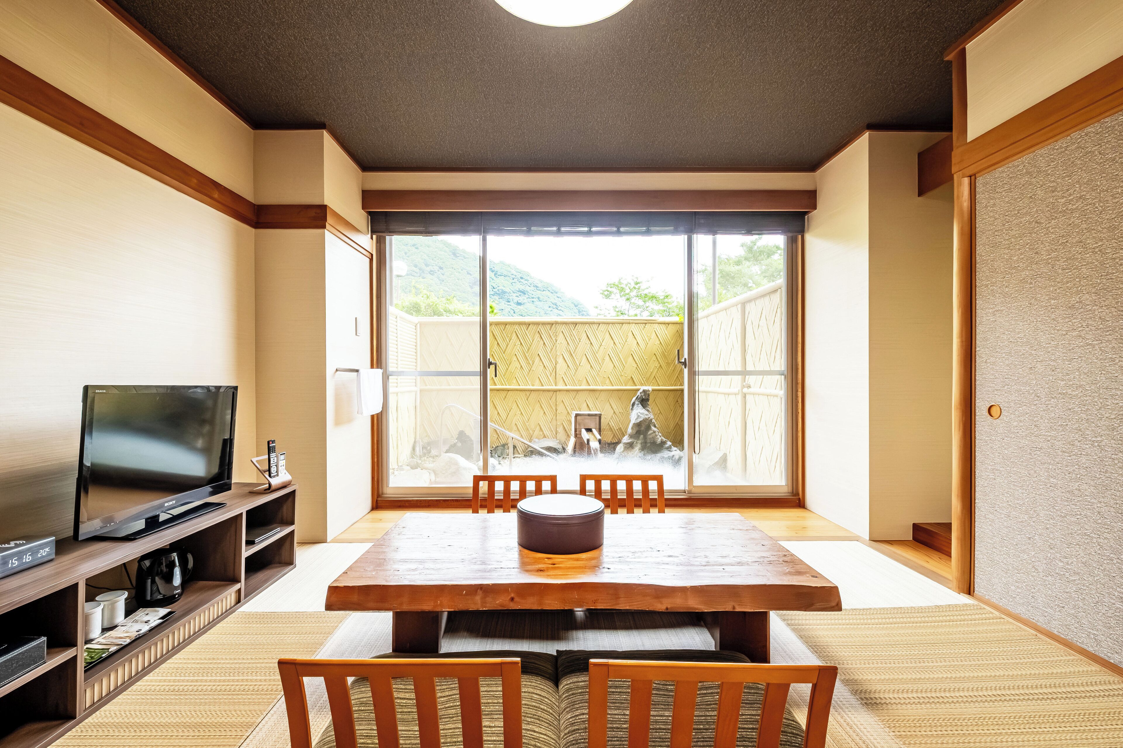 Una habitación tradicional japonesa con suelo de tatami, una mesa de madera baja con cojines, un televisor sobre un soporte de madera y una puerta corredera de cristal que da a un espacio exterior con vistas a la vegetación.