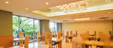 A spacious dining area with wooden tables and chairs, large windows offering a view of trees, and recessed lighting on the ceiling.