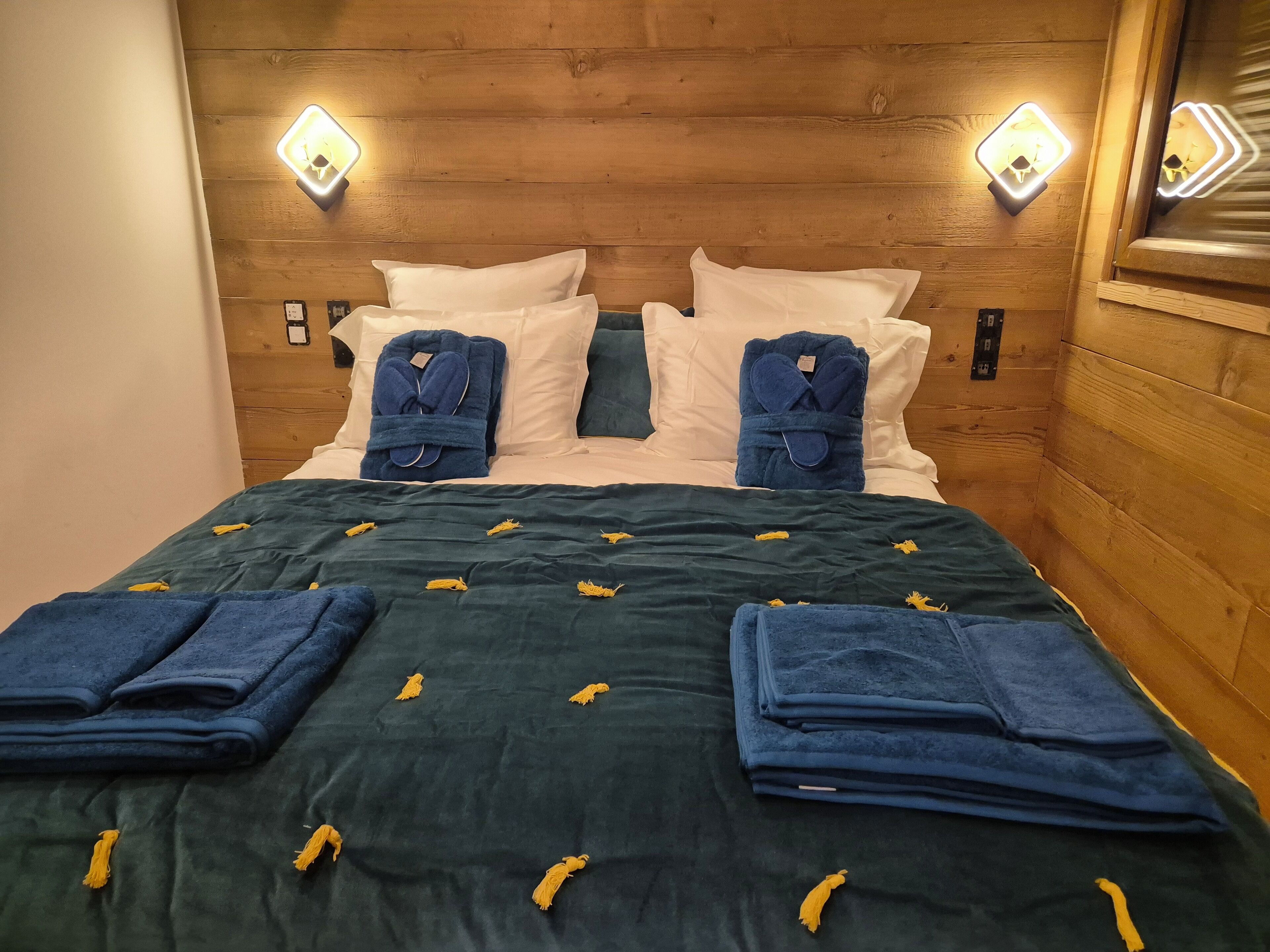 A hotel room with a wooden headboard, two wall-mounted lights, white and blue pillows, a green bedspread with yellow tassels, and folded blue towels.