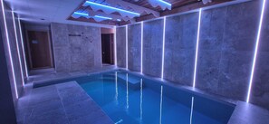 Una piscina interior moderna con superficie de agua azul cristalina, rodeada de paredes de piedra e iluminada por luces LED azules.