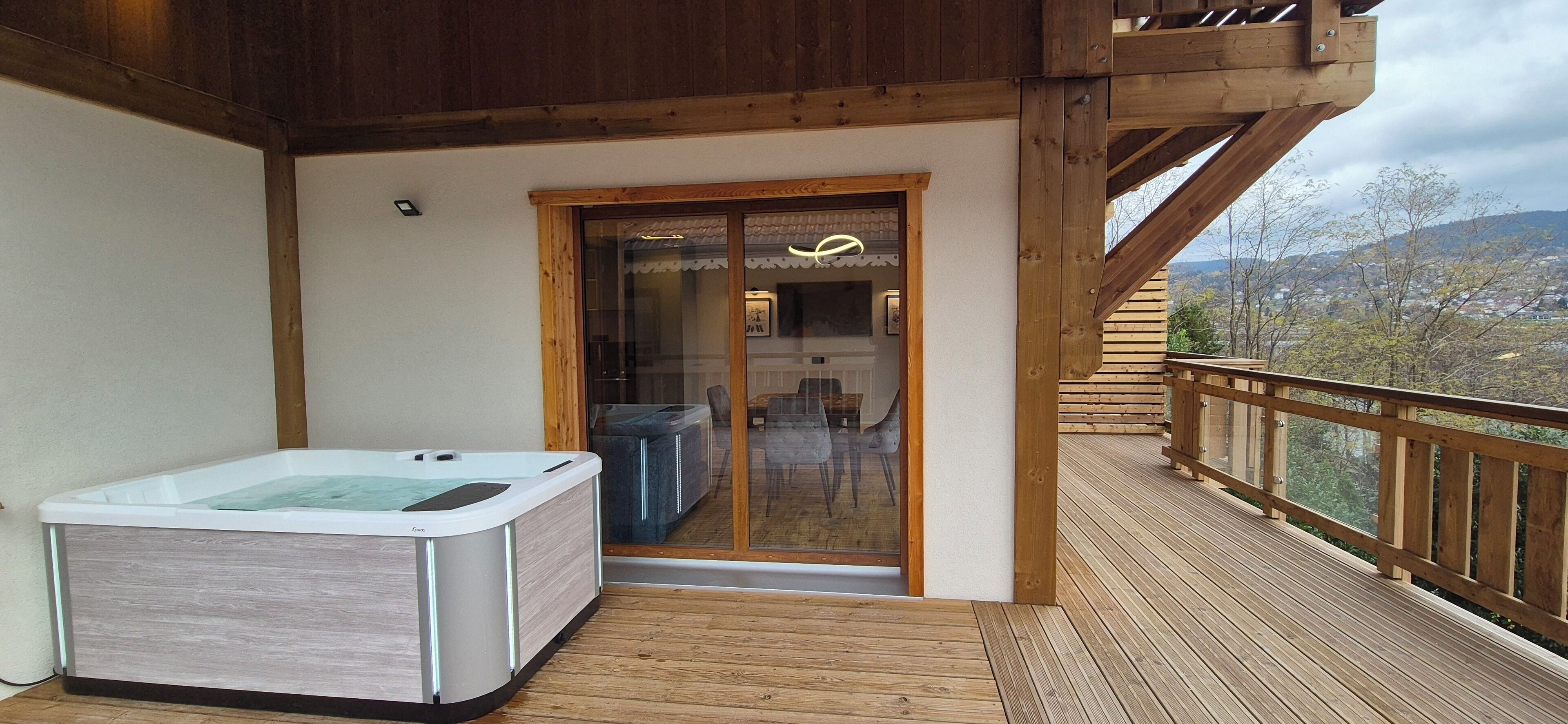 A wooden deck with a hot tub and a glass door leading to an interior space with a table and chairs.
