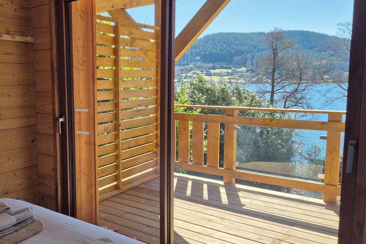 A wooden cabin room with a bed, a wooden door, and a balcony with a view of a lake and mountains.