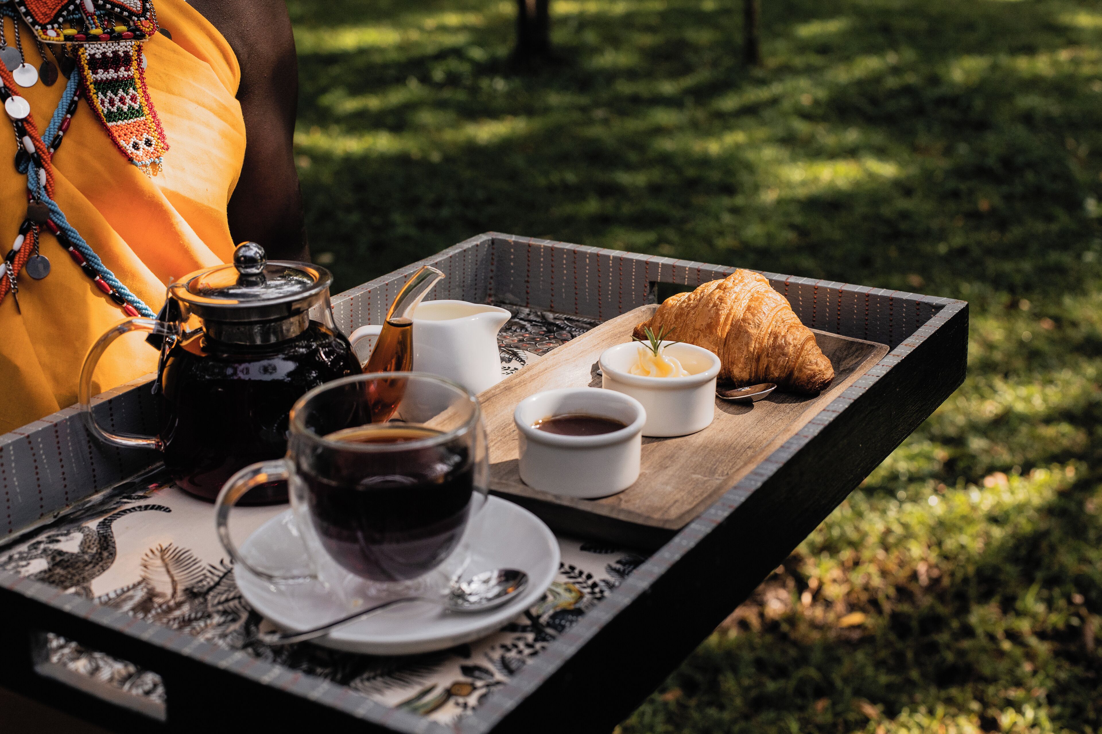 Een dienblad met een croissant, koffie en een theepot op een tafel buiten.