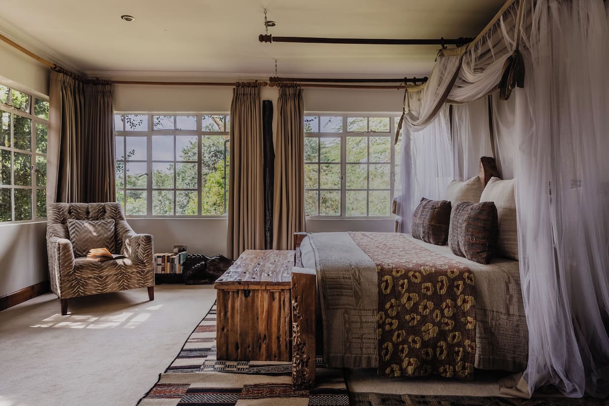 A bedroom with a bed, a chair, a wooden table, and large windows with curtains.