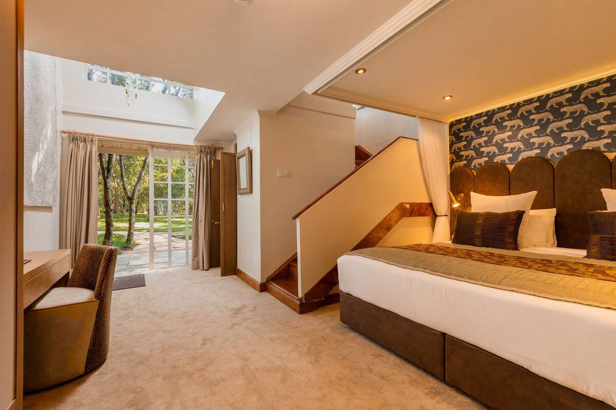 A bedroom with a large bed, a desk, a chair, a staircase, and a view of the outdoors.
