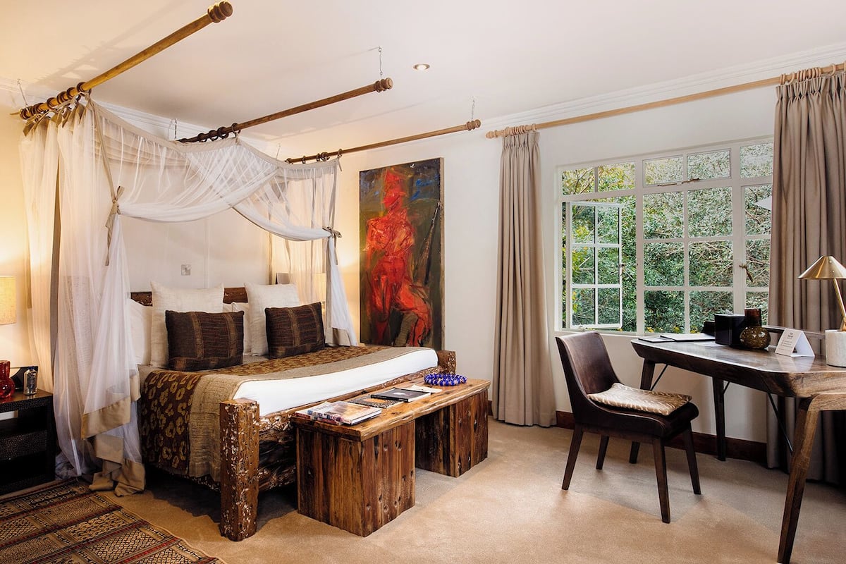 A bedroom with a canopy bed, a desk with a chair, a large window, and a painting on the wall.