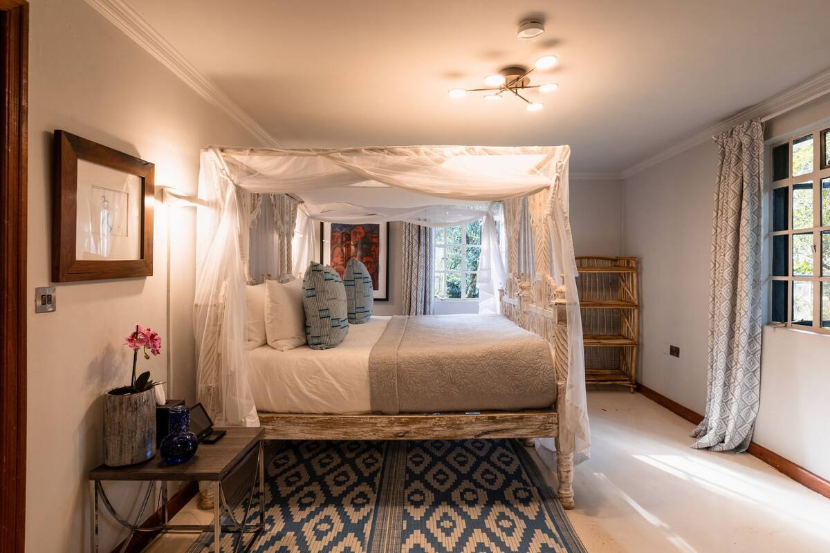 A bedroom with a canopy bed, a bedside table with a vase and a laptop, a patterned rug, and a window with curtains.