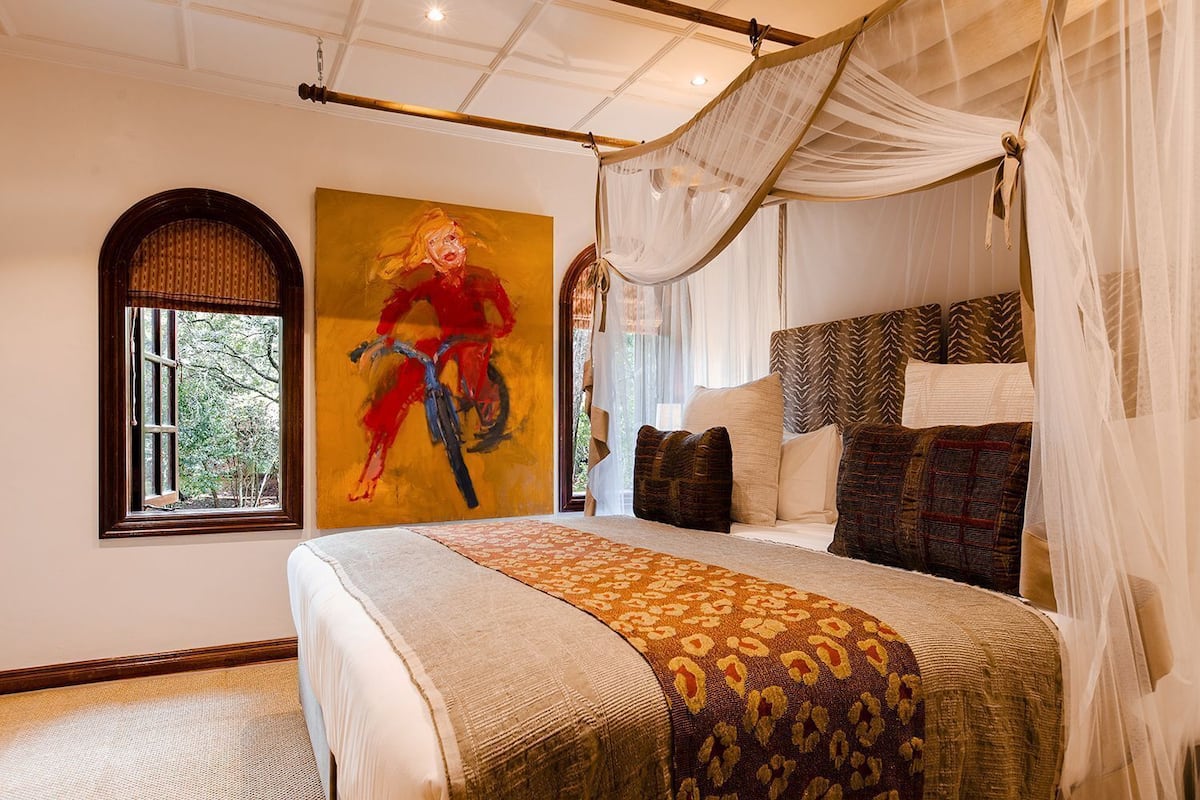 A bedroom with a canopy bed, a large painting, and a window with a view of trees.