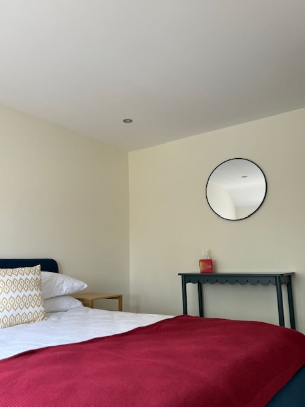 Una cama bien hecha con un edredón rojo, una mesita de noche con un objeto rojo y un espejo redondo en la pared.