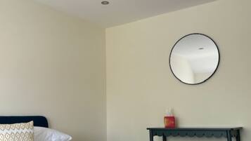 Una cama bien hecha con un edredón rojo, una mesita de noche con un objeto rojo y un espejo redondo en la pared.