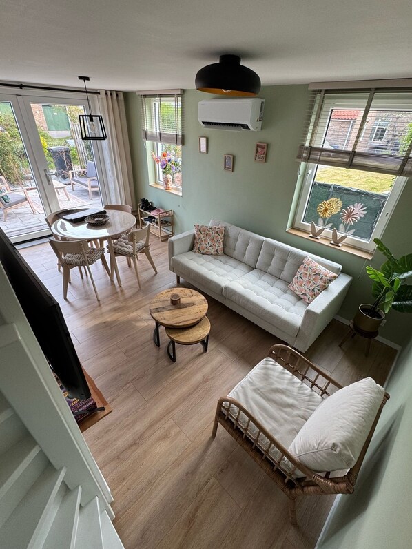 A spacious living room with a sofa, a round wooden coffee table, and a wicker chair. There is a dining table with chairs, a large window with blinds, and an air conditioning unit mounted on the wall.