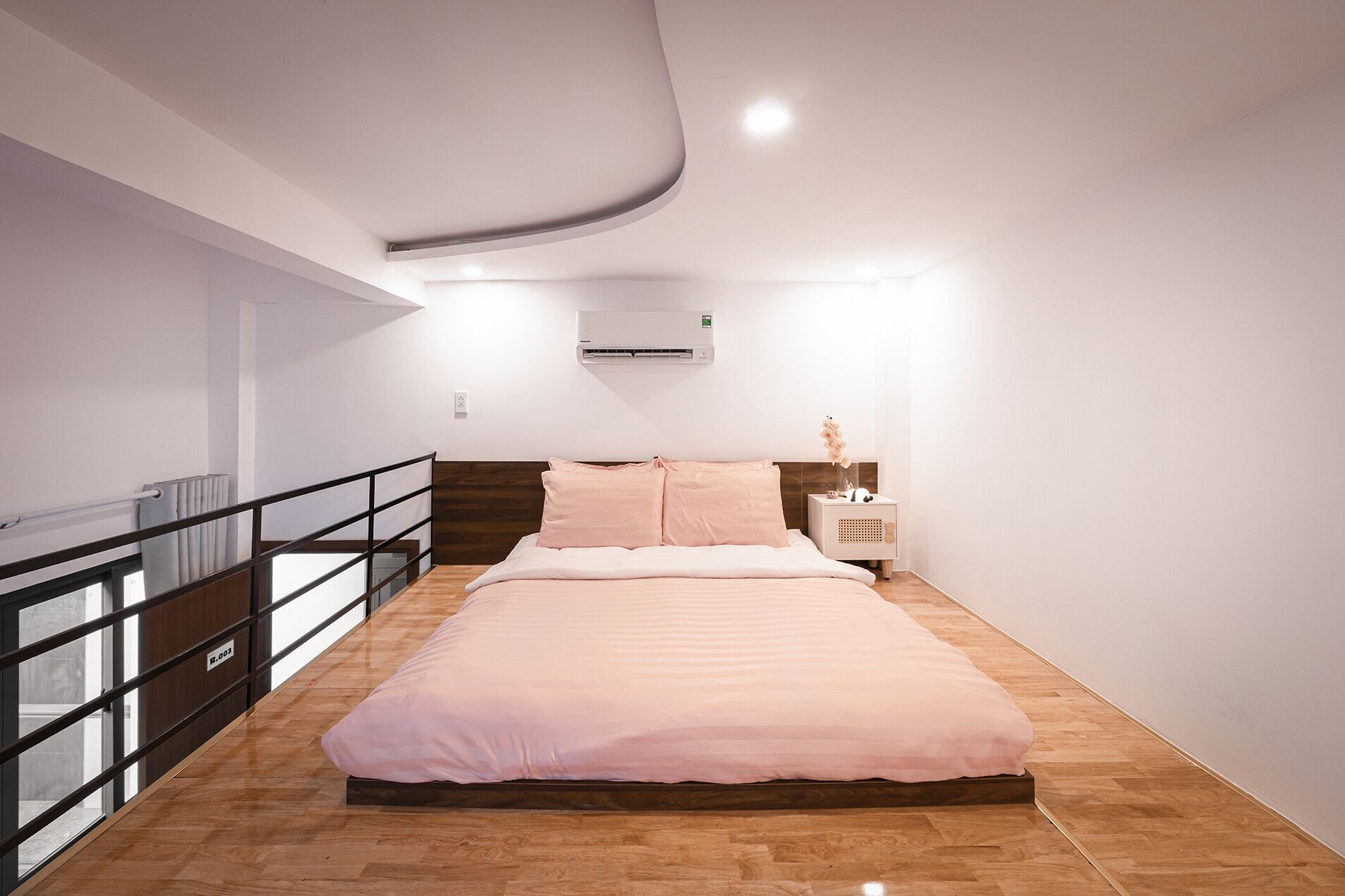 A modern bedroom with a large bed, a nightstand, an air conditioning unit, and a balcony with a railing.