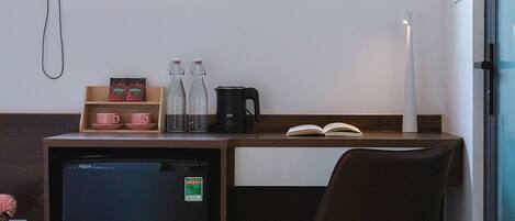 A compact hotel room with a small refrigerator, a desk with a chair, a bedside table with a book and a lamp, and a window with a view of buildings.