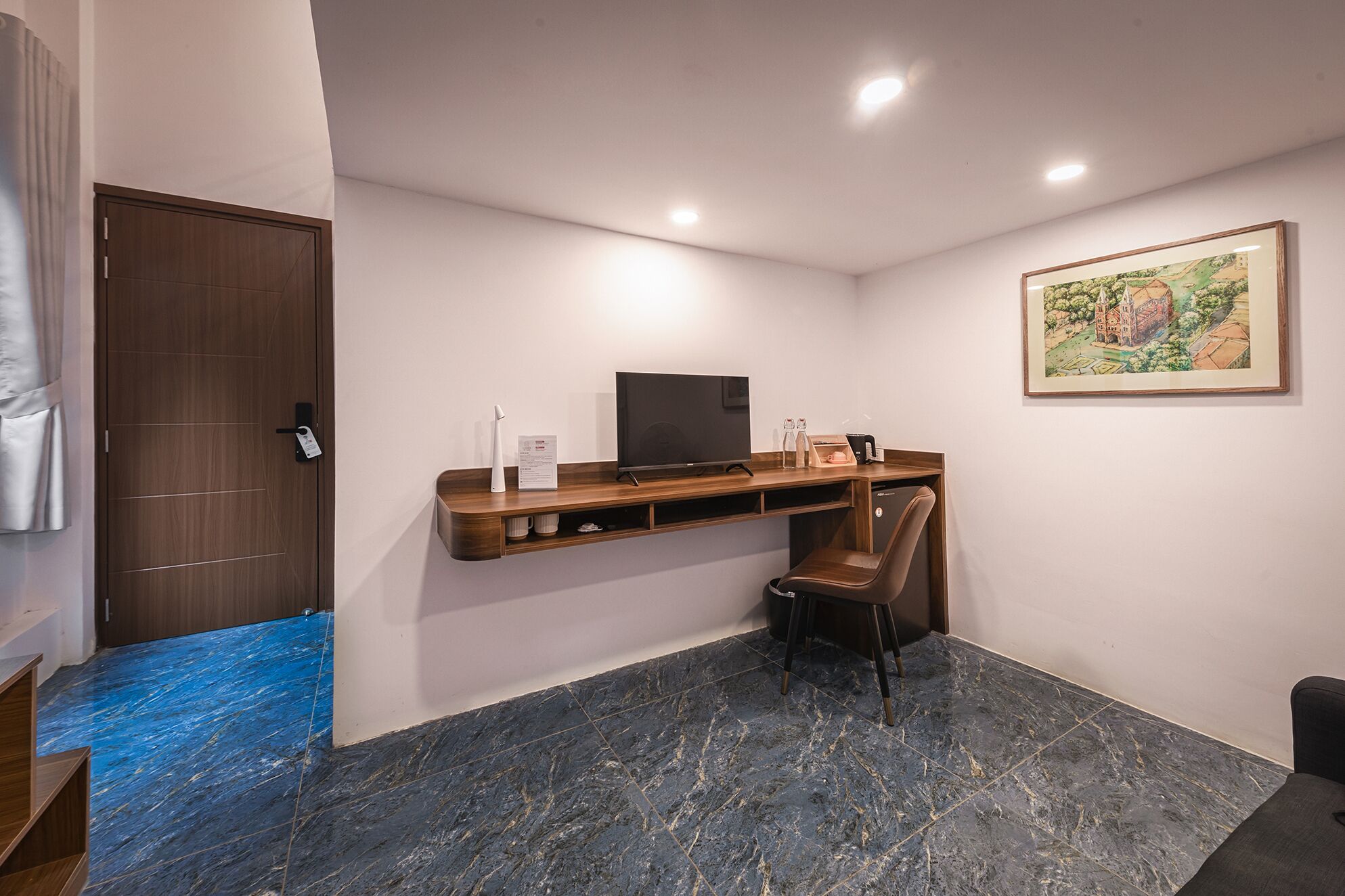 A modern hotel room with a dark marble floor, a wooden desk with a flat-screen TV, a chair, and a framed painting on the wall.