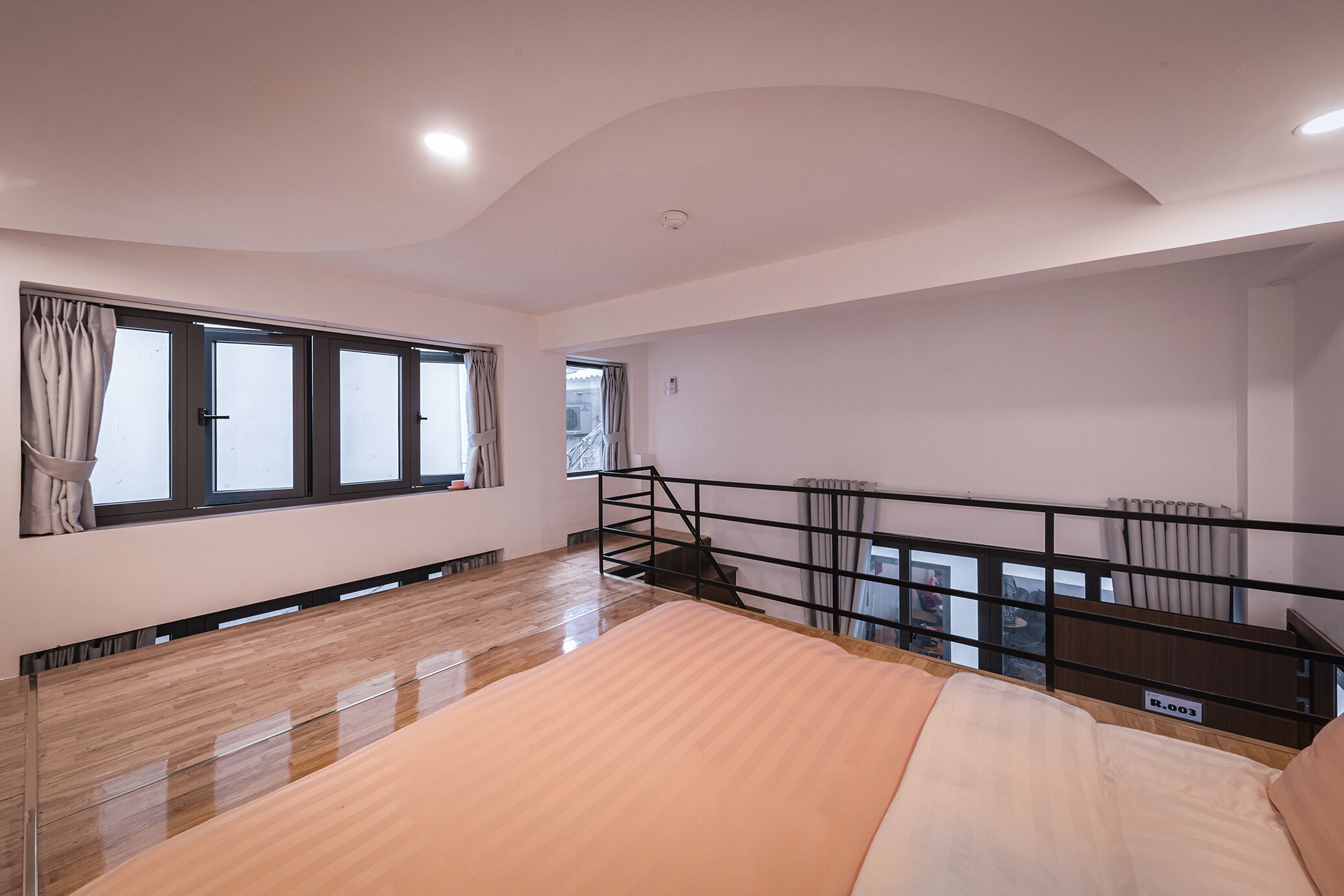 A spacious room with a large bed, a wooden floor, a balcony with a railing, and a window with curtains.
