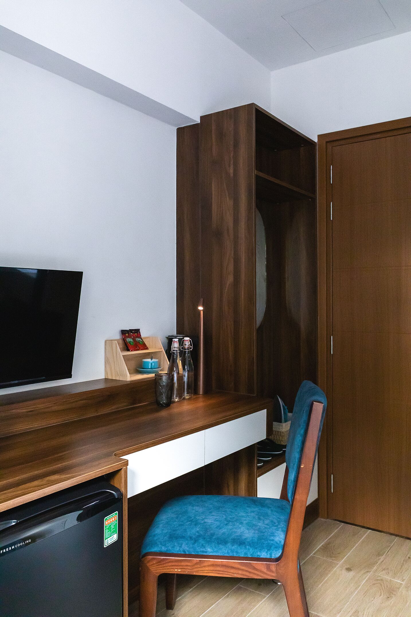 A compact room with a wooden desk, a television, a small refrigerator, a blue chair, and a wardrobe.
