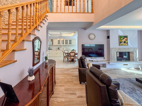 A spacious living room with a wooden staircase, a dining area, and a kitchen in the background. - Villa Zeytin (Kusadasi)