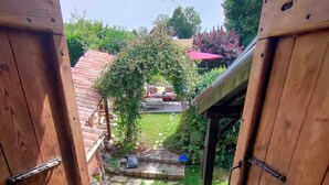 Ein hölzernes Tor führt in einen Garten mit einem kleinen Tisch und Stühlen, einer Pergola und einem Gebäude mit rotem Dach im Hintergrund.