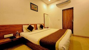 A hotel room with a wooden headboard, a bed with white and brown bedding, a bedside table with a water bottle, a mirror, and a wooden door. - Aangan Palace (Udaipur)