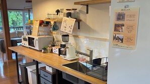 A kitchen with a wooden countertop, a coffee machine, a kettle, and a refrigerator. There is a menu hanging on the wall and a window with a view of trees. - Private Room Guesthouse ORSA MINAMIASO (Minamiaso)