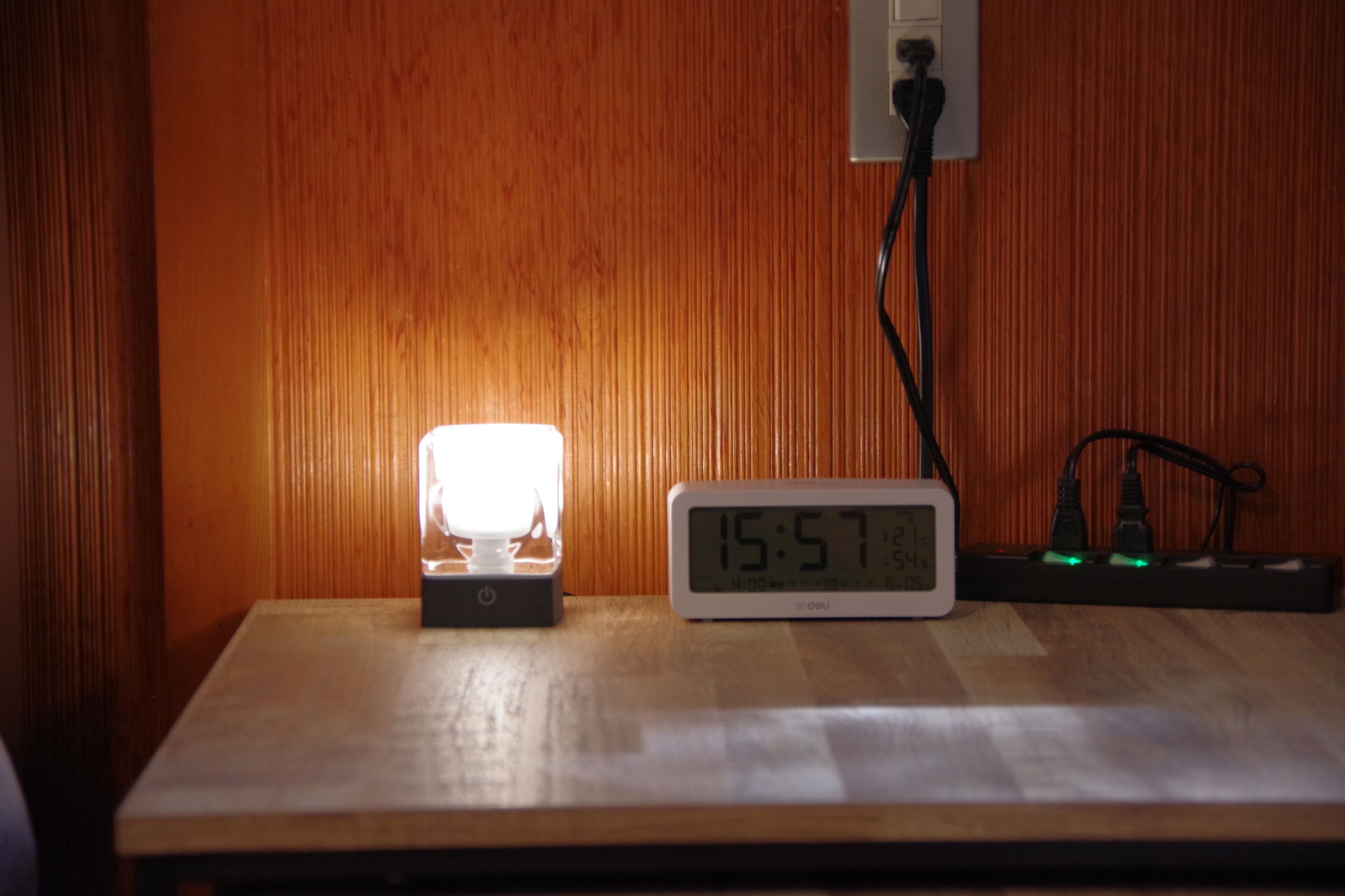 Uma mesa de madeira com um relógio digital mostrando 15:57 e uma pequena luminária.