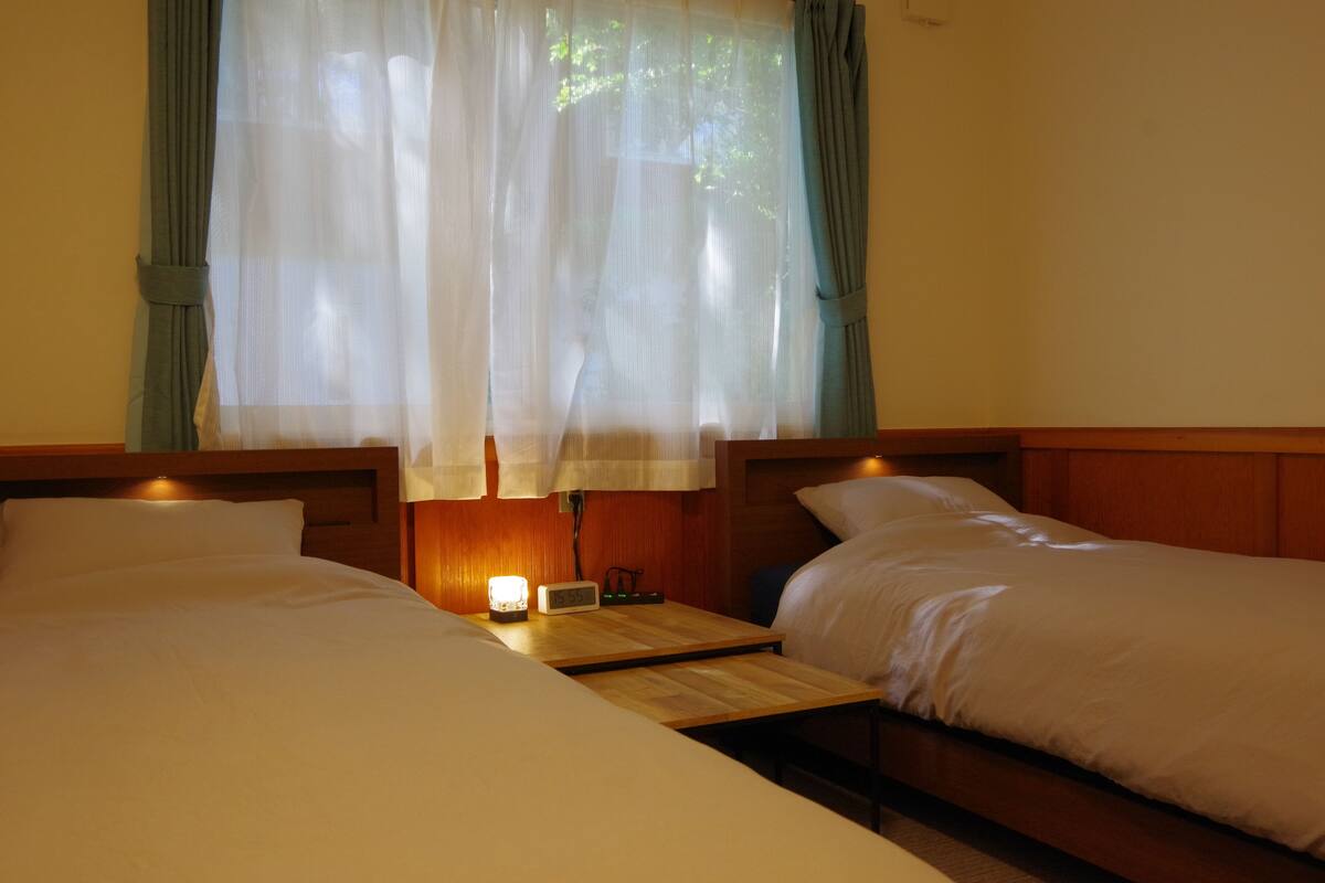 A room with two beds, a nightstand with a lamp, and sheer curtains.