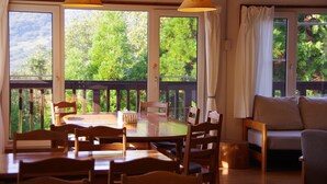 A dining area with a round table and chairs, large windows with curtains, and a view of greenery outside. - Private Room Guesthouse ORSA MINAMIASO (Minamiaso)