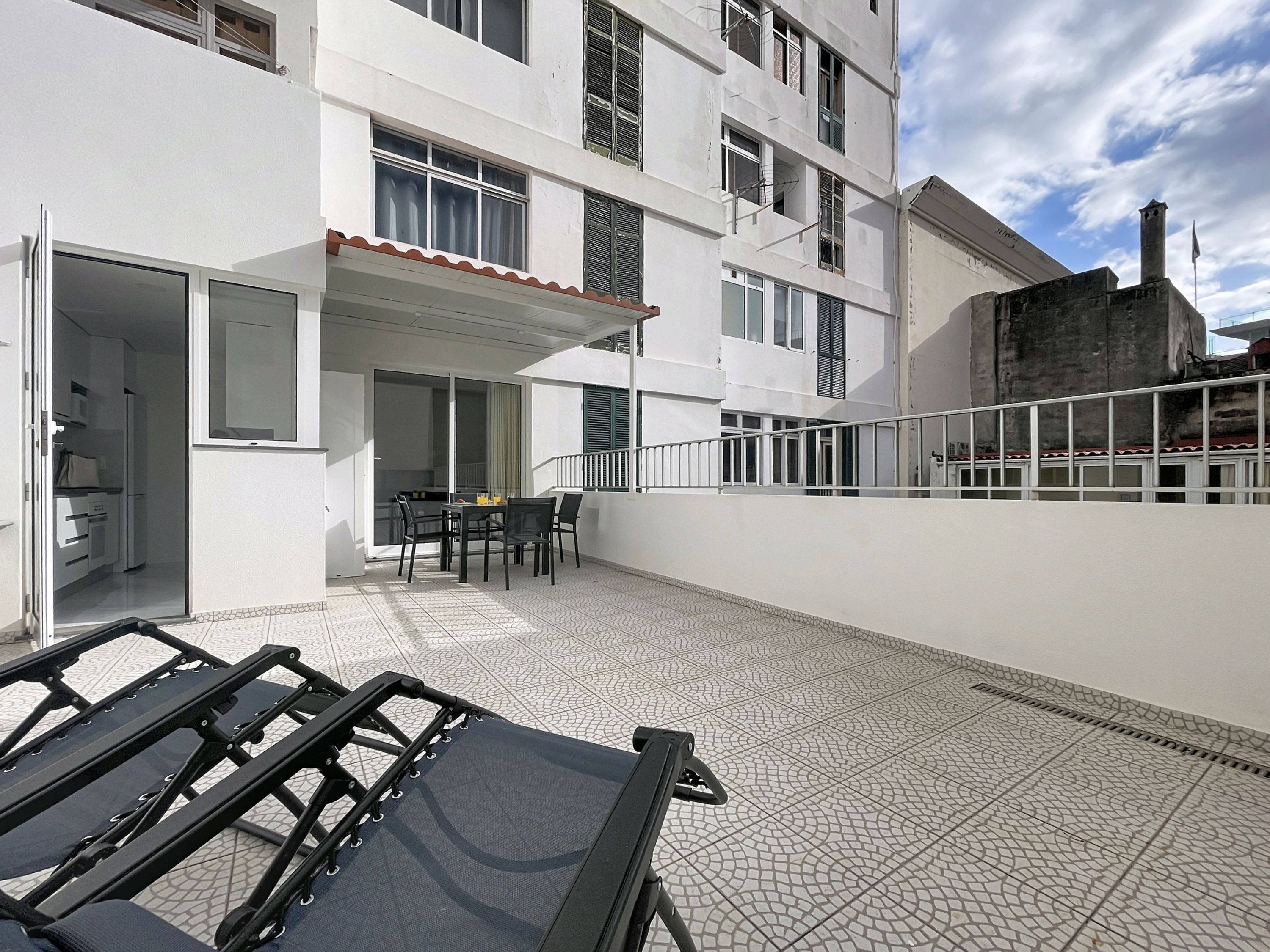 A balcony with a table and chairs, a sliding glass door leading to a kitchen, and a white building with multiple windows and balconies.