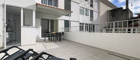 A balcony with a table and chairs, a sliding glass door leading to a kitchen, and a white building with multiple windows and balconies.