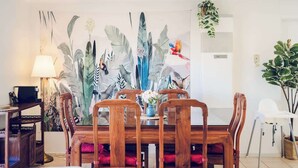 A dining area with a wooden table and chairs, a wall mural with tropical leaves, a lamp, and a potted plant. - Entire Cozy 2-Bedroom House in Mount Waverley (Mount Waverley)