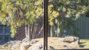 A window view of a fenced outdoor area with trees, a brick wall, and a wooden fence. - Entire Cozy 2-Bedroom House in Mount Waverley (Mount Waverley)