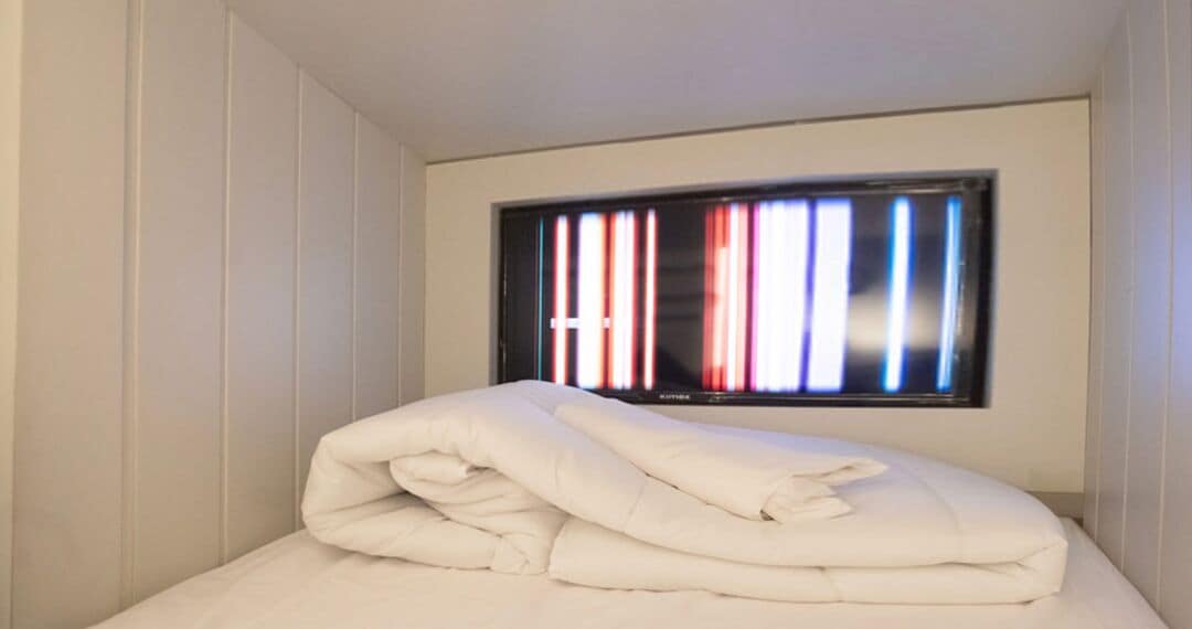 A neatly made bed with a white comforter and pillows, a window with vertical blinds, and white walls.