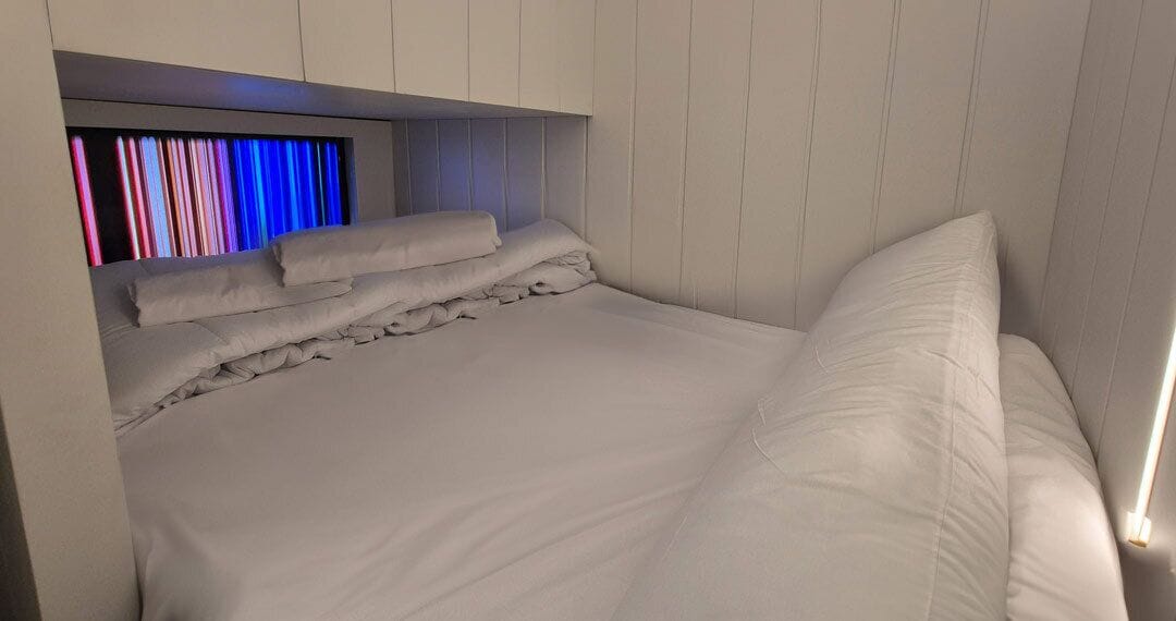 Uma cama bem arrumada com lençóis e almofadas brancas, cabeceira com iluminação embutida e teto inclinado.